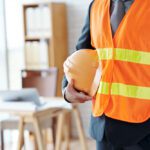 Person in a high-visibility orange safety vest holding a hard hat indoors, likely a site supervisor in an office setting.