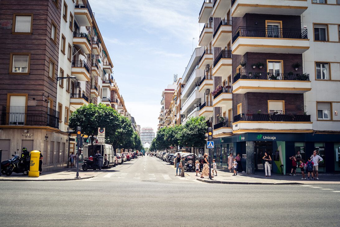 781457 Fincas Legio | diario-economia.com Sunlit city street lined with multi-story buildings, pedestrians crossing at a crosswalk and cars parked along both sides.