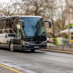 Black tour bus driving through a city street with blurred storefronts in the background, reflections on its glossy surface.