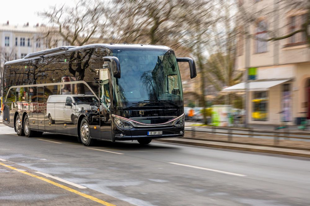 Black tour bus driving through a city street with blurred storefronts in the background, reflections on its glossy surface.
