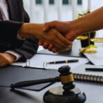 Two professionals in business attire shake hands across a desk, with a gavel, notebook, and laptop nearby, suggesting a legal agreement.