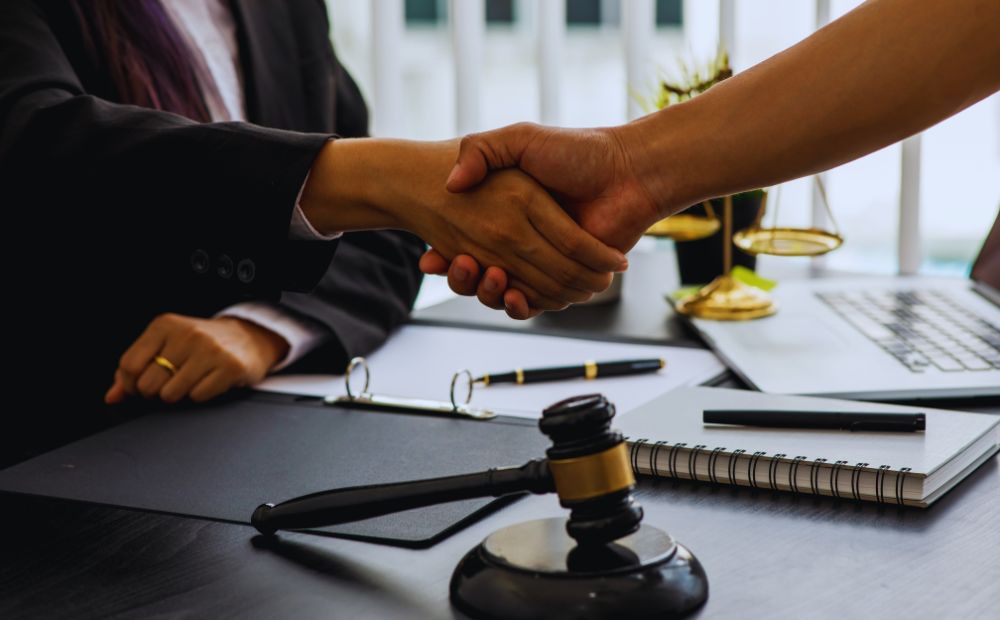 782214 - Grupo AB Abogados | diario-economia.com Two professionals in business attire shake hands across a desk, with a gavel, notebook, and laptop nearby, suggesting a legal agreement.