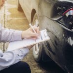 Man in a checkered shirt writes on a clipboard beside a damaged black car on the street
