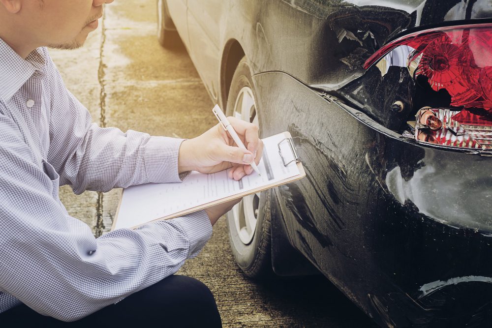 782226 - Zeeguros | diario-economia.com Man in a checkered shirt writes on a clipboard beside a damaged black car on the street
