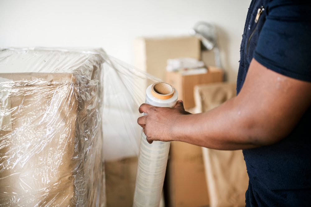782494 - Mudanzas Castilla | diario-economia.com Person wrapping a pallet with clear plastic wrap using packing tape in a storage area with cardboard boxes in the background