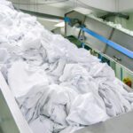 Industrial laundry conveyor dumping a large pile of white linens into a processing line, in a factory setting.