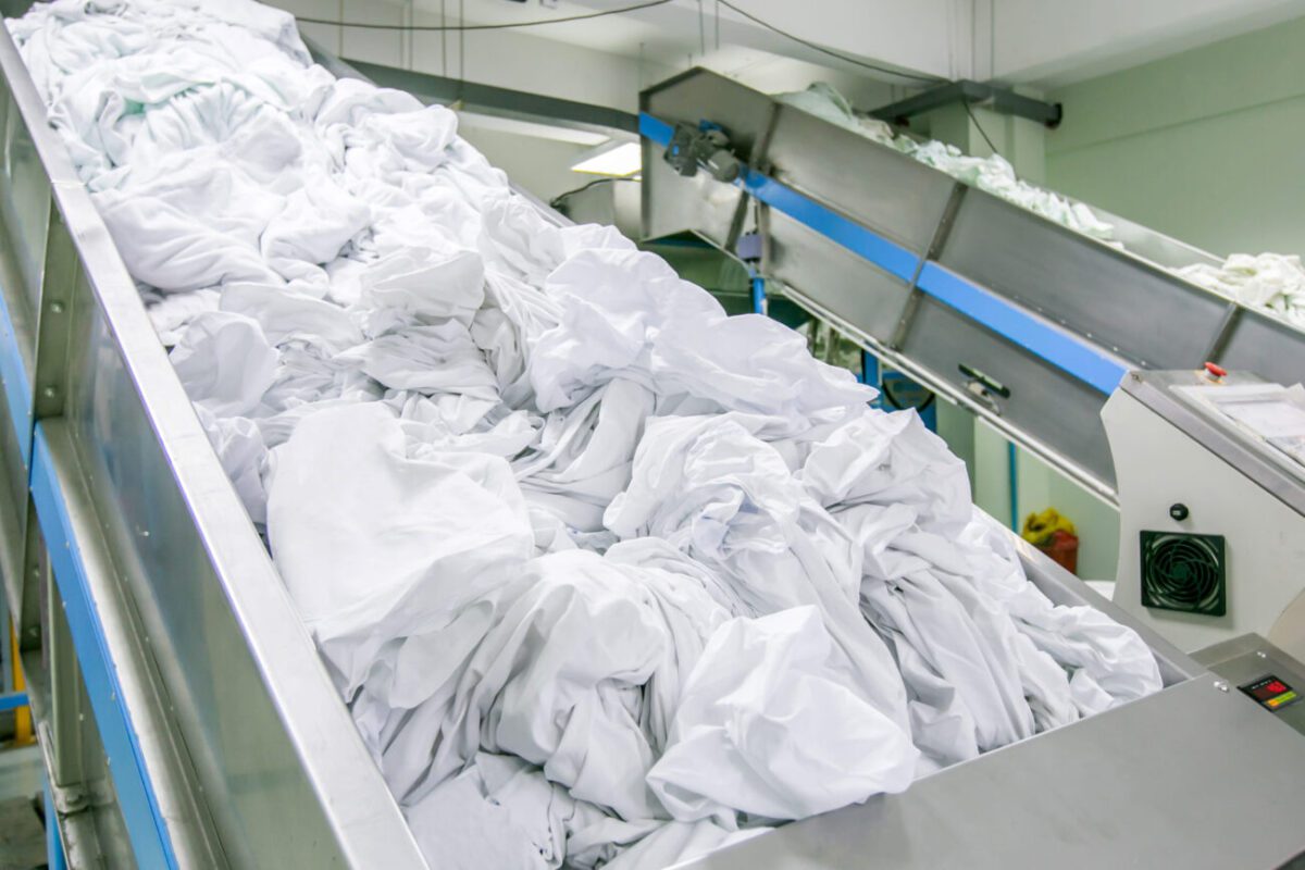 Industrial laundry conveyor dumping a large pile of white linens into a processing line, in a factory setting.