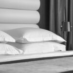 Black-and-white photo of a bed with white pillows, a padded headboard, and a standing lamp by dark curtains.