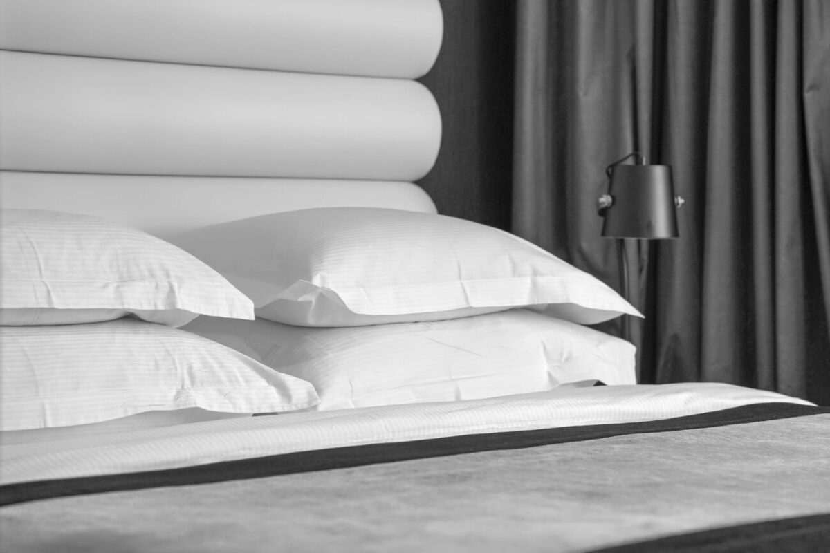 Black-and-white photo of a bed with white pillows, a padded headboard, and a standing lamp by dark curtains.