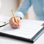 Person writes on a form with a pencil on a desk; eyeglasses rest nearby on the table.