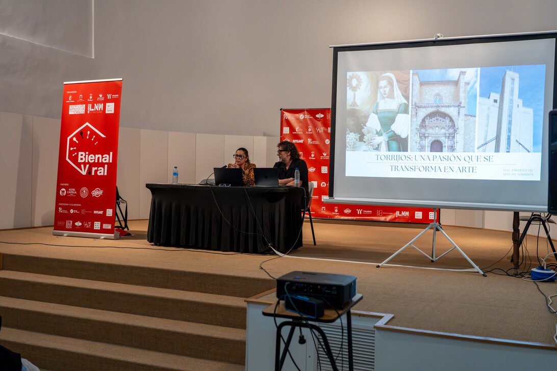 Bienal viral - IP | diario-economia.com Two speakers sit at a table on a stage, with a projector screen displaying a slide about Torrijos and art in the background.