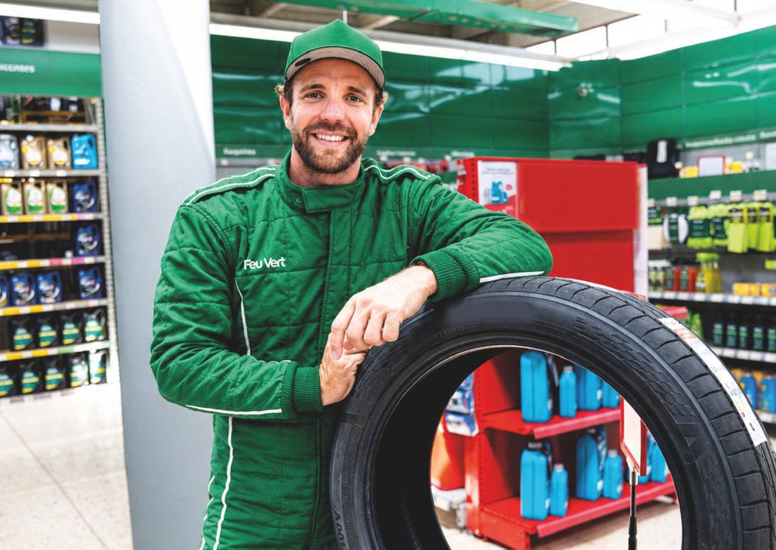 Dani Clos - Feu Vert - IP | diario-economia.com Smiling male mechanic in a green uniform leaning on a car tire in a store aisle.
