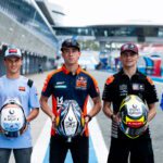Five race car drivers stand in a pit lane, each holding a helmet and wearing team uniforms with sponsor logos.