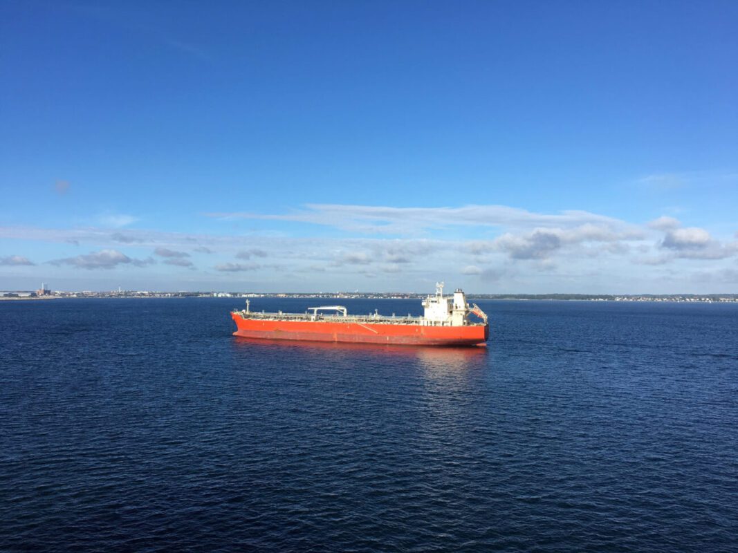 Large orange tanker sailing on calm blue sea with a distant shoreline under a clear sky, white superstructure toward the stern.