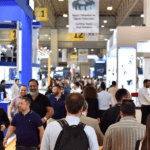 Busy trade show floor with attendees walking between booths, display stands, and hanging banners in a large exhibition hall.