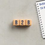 Wooden blocks spell B2B beside a notebook that says 'Business to Business' with a silver pen nearby, on a light surface.