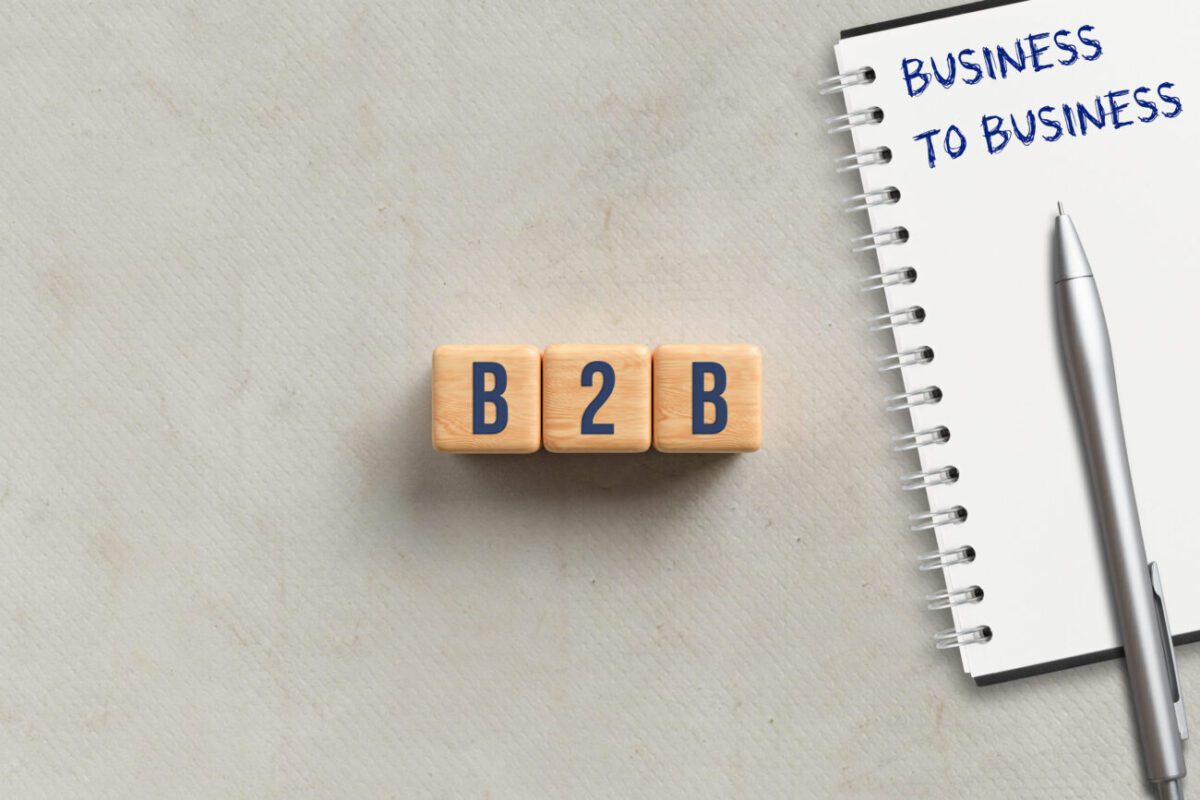 Wooden blocks spell B2B beside a notebook that says 'Business to Business' with a silver pen nearby, on a light surface.