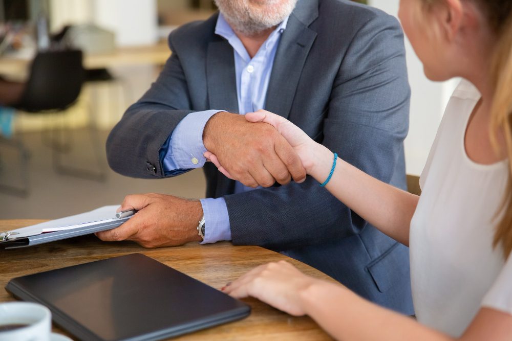 778552 - CGS Abogados | diario-economia.com Two professionals shake hands over a wooden table in a business meeting, with documents and a tablet nearby cheery room in background.