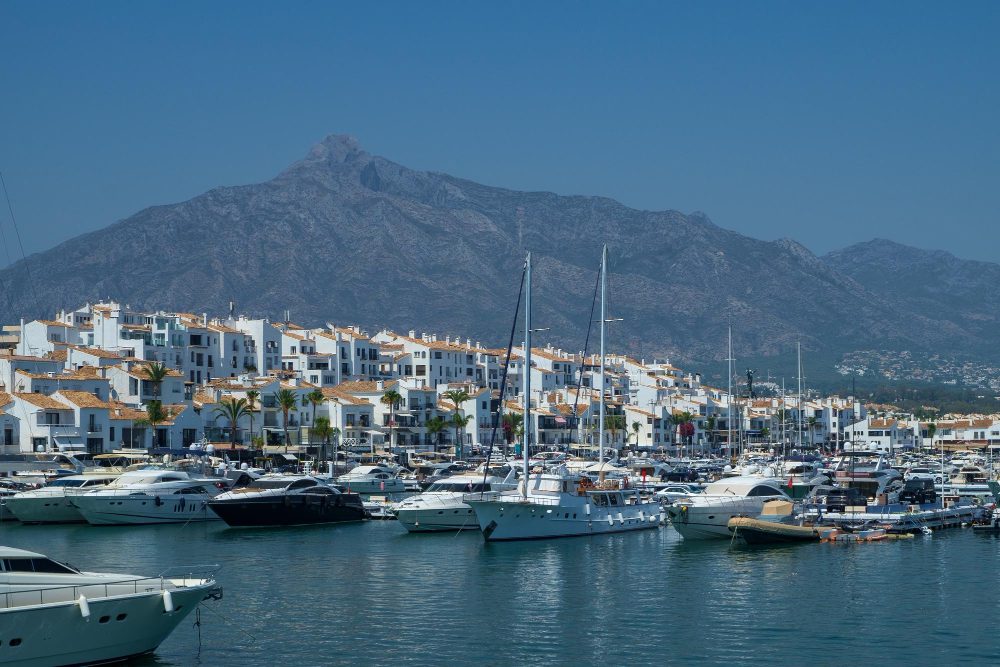 780234 - Absolute Prestige | diario-economia.com Marina with numerous white yachts docked, backed by white buildings with orange-tiled roofs and a mountain range beyond.