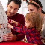 Family of three shares a Christmas moment, placing treats from a white Advent calendar together at a table with a red cloth, smiling faces.