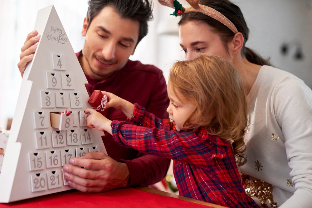 780628 - Loterías Doña Sol | diario-economia.com Family of three shares a Christmas moment, placing treats from a white Advent calendar together at a table with a red cloth, smiling faces.