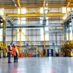 Two workers in safety vests review plans inside a large industrial maintenance hall with yellow overhead cranes and heavy equipment nearby.