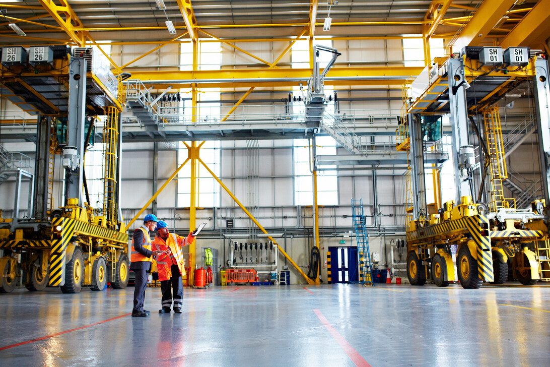 Men tablet and discussion in warehouse for shipping distribution schedule or planning export Workers tech and talking in storage space for logistics management import control or expansion ideas | diario-economia.com Two workers in safety vests review plans inside a large industrial maintenance hall with yellow overhead cranes and heavy equipment nearby.