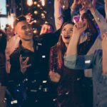 Group of smiling friends dancing at a lively party with confetti and warm lights.
