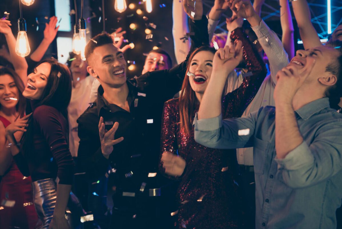Group of smiling friends dancing at a lively party with confetti and warm lights.