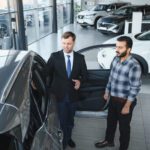 Salesperson in a suit showing a silver car with an open door to a customer inside a bright showroom with other cars in the background.