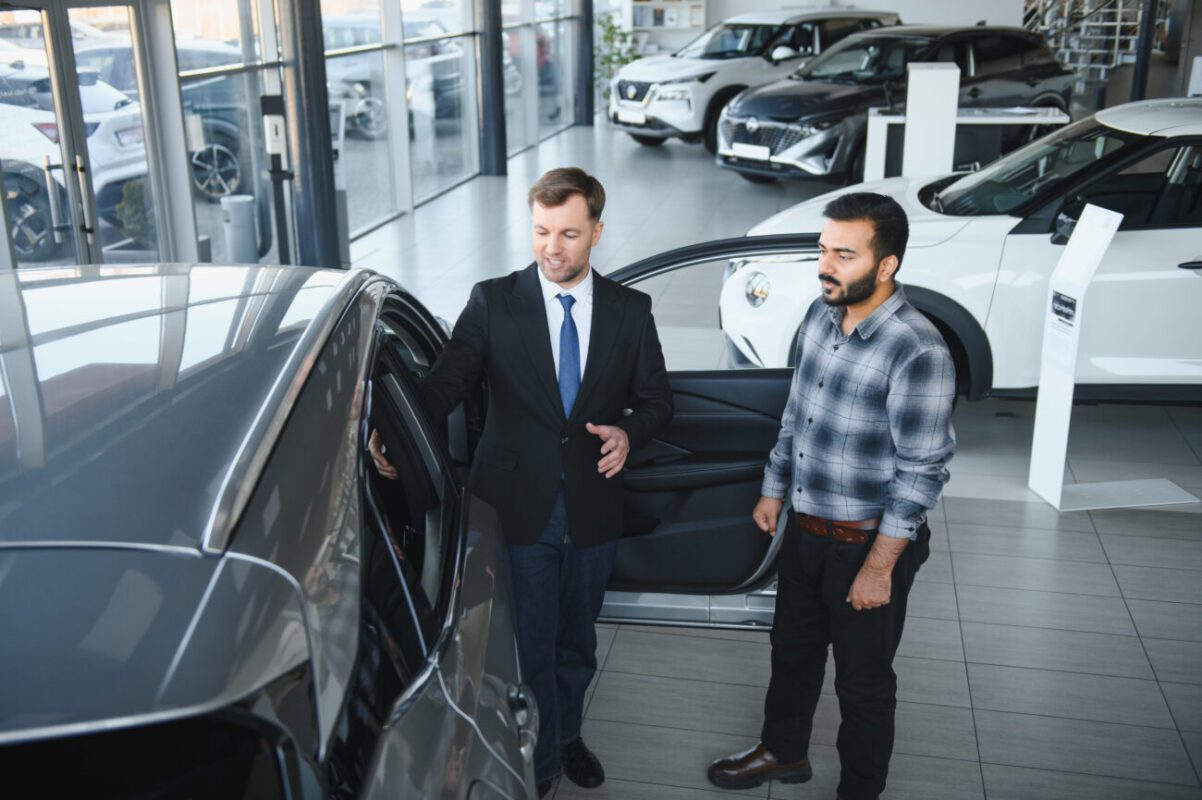 784317 Grupo CMR | diario-economia.com Salesperson in a suit showing a silver car with an open door to a customer inside a bright showroom with other cars in the background.