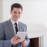 Smiling man in a gray suit holding a tablet in a bright room near a fireplace.