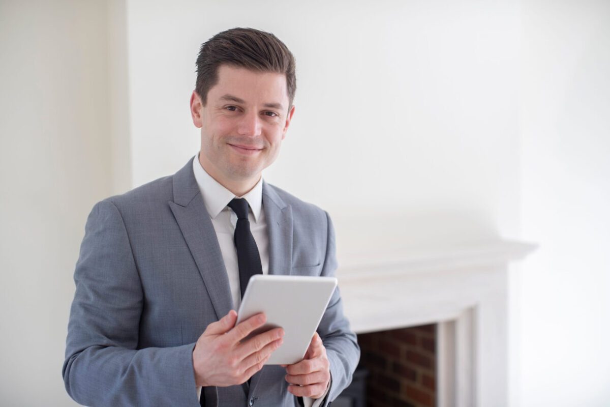 Smiling man in a gray suit holding a tablet in a bright room near a fireplace.