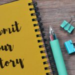 Yellow spiral notebook page reads 'Submit Your Story' beside a mint pen and blue binder clips on a wooden desk surface.