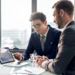 Two professionals in suits discuss charts on a laptop screen at a bright office meeting by a glass table.