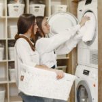 Two women at a laundromat load a front-loading washer, one places a towel inside while the other holds a decorative white laundry basket.
