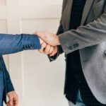 Two professionals shake hands in a business agreement; one wearing a blue plaid suit, the other a gray blazer.