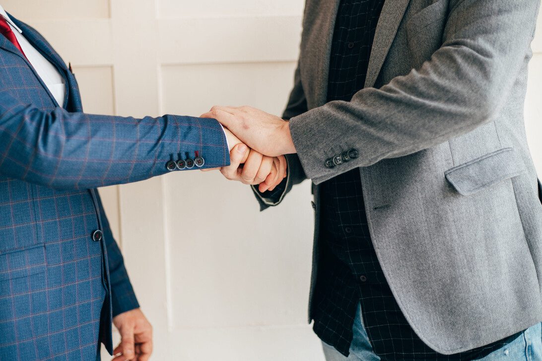 Business people handshake success teamwork | diario-economia.com Two professionals shake hands in a business agreement; one wearing a blue plaid suit, the other a gray blazer.