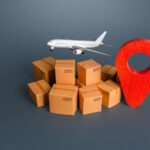 Model airplane flying over a pile of cardboard boxes and a large red map pin, symbolizing air shipping to a destination.