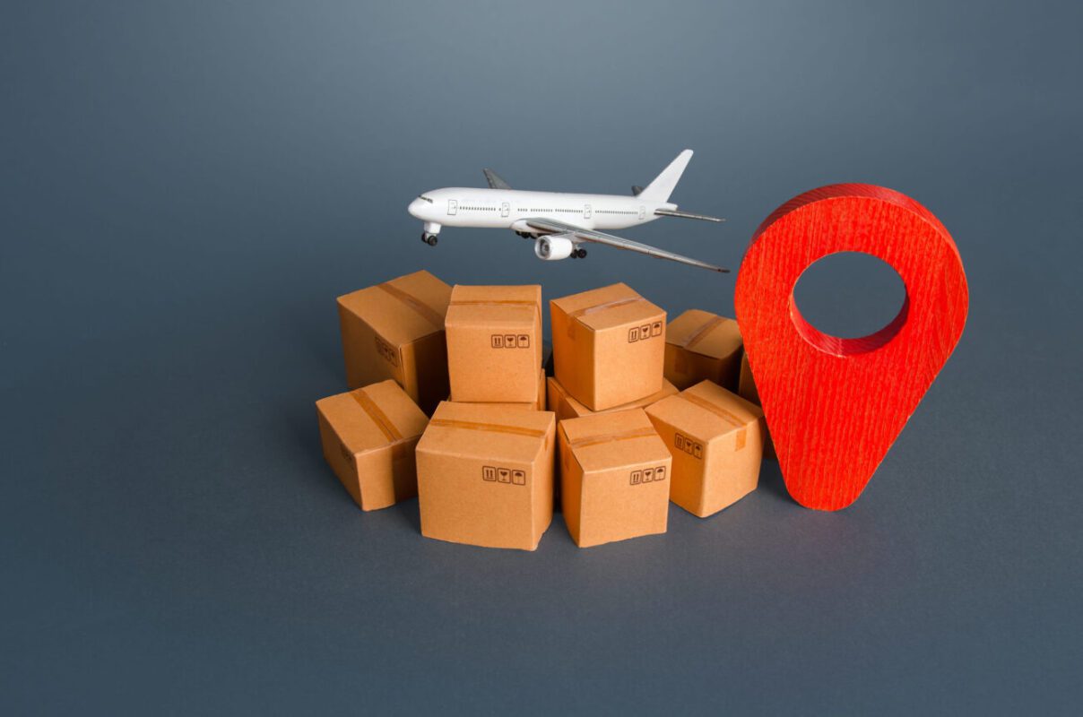 785100 IAS Handling | diario-economia.com Model airplane flying over a pile of cardboard boxes and a large red map pin, symbolizing air shipping to a destination.