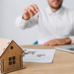 Man in a white shirt extends a key toward a small wooden house model on a desk, with a laptop and papers in the background — real estate scene.