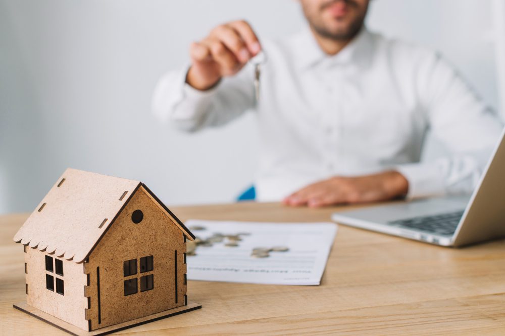 785139 - Soms Granados | diario-economia.com Man in a white shirt extends a key toward a small wooden house model on a desk, with a laptop and papers in the background — real estate scene.