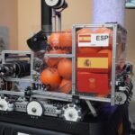 Robotic machine on display with orange balls, ESP label, and Spain flag stickers inside a metal frame.