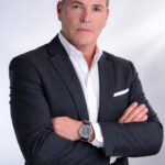 Professional man in a navy suit with white shirt, arms crossed, neutral expression, business portrait.