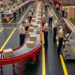 Warehouse workers line a red curved conveyor belt, packing and labeling cardboard boxes in a large facility.