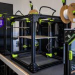 Open-frame 3D printer with neon-green clamps and spool reels on top, showing a flat build platform inside a glass enclosure.